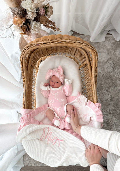 Newborn baby in a pink outfit lying in a wicker crib with decorative flowers in the background.