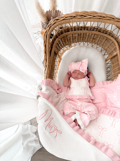 Baby in white and pink singlet top, pink and white pants, on a bow print blanket, in a wicker basket with dried florals in the background and sheer curtains surrounding two sides. 