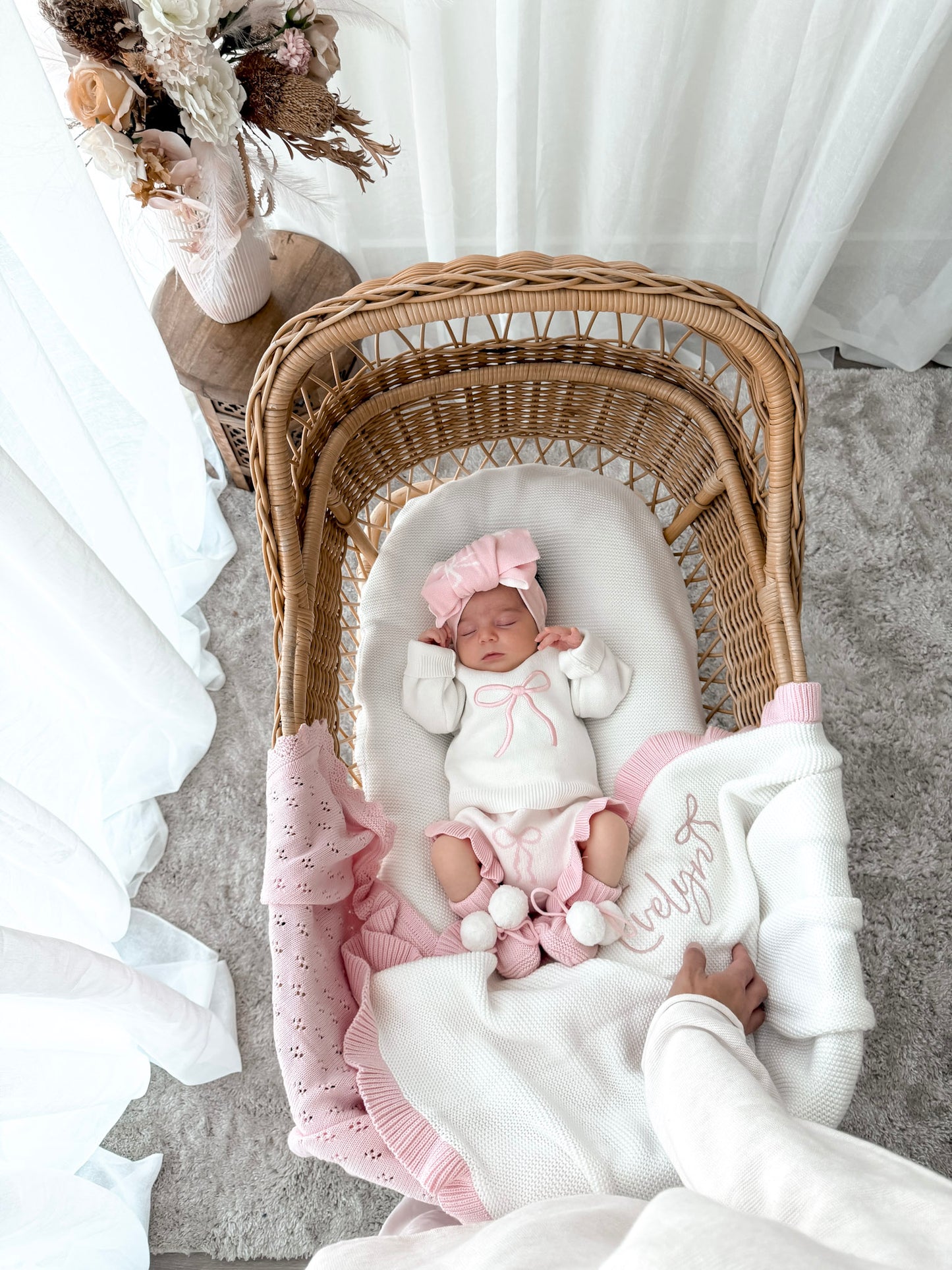 Newborn wearing a white jumper with a pink bow, bloomer bottoms with bow pattern and frills, bow pattern top knot, with a personalised whtie and pink blanket, in a wicker basket, sheer curtains and decorative dried florals.