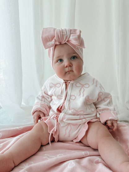 Baby wearing a pink outfit with a bow headband against a white curtain background