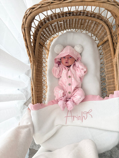 Baby in a pink hooded outfit with pom poms lying in a wicker crib with a white blanket. 
