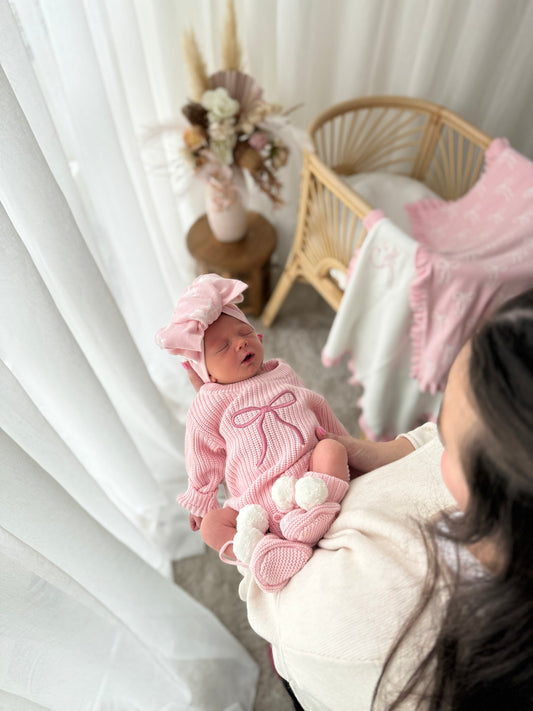 Mother carrying her baby on her forearm, baby laying asleep in a pink outfit with a dark pink bow romper, bow headback and booties. 