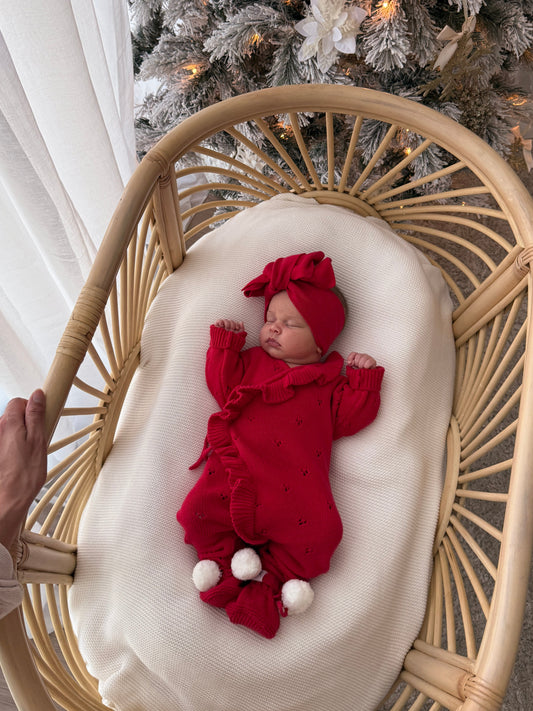A newborn baby wearing a red knit outfit and matching hat is lying on a white cushion inside a woven rattan bassinet. A hand is holding the bassinet, and a decorated Christmas tree is visible in the background.