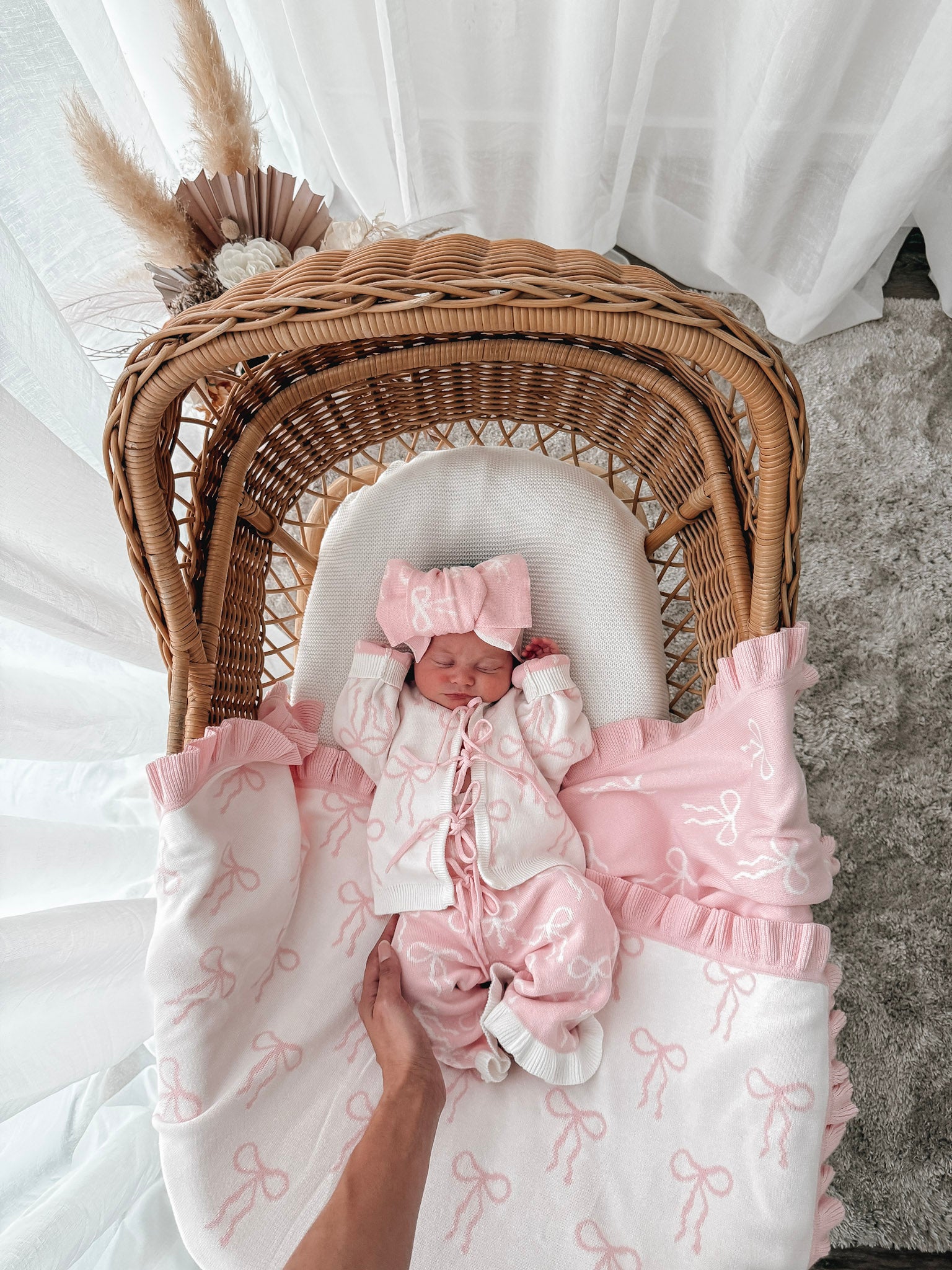 Newborn baby in a bow print cardigan, that ties in the front,bow print flared pants, both in pink & white, with a matching top knot. Baby is in a wicker backet, decorative dried florals behind. 