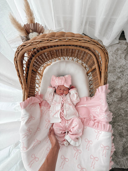 Newborn baby in a bow print cardigan, that ties in the front,bow print flared pants, both in pink & white, with a matching top knot. Baby is in a wicker backet, decorative dried florals behind. 