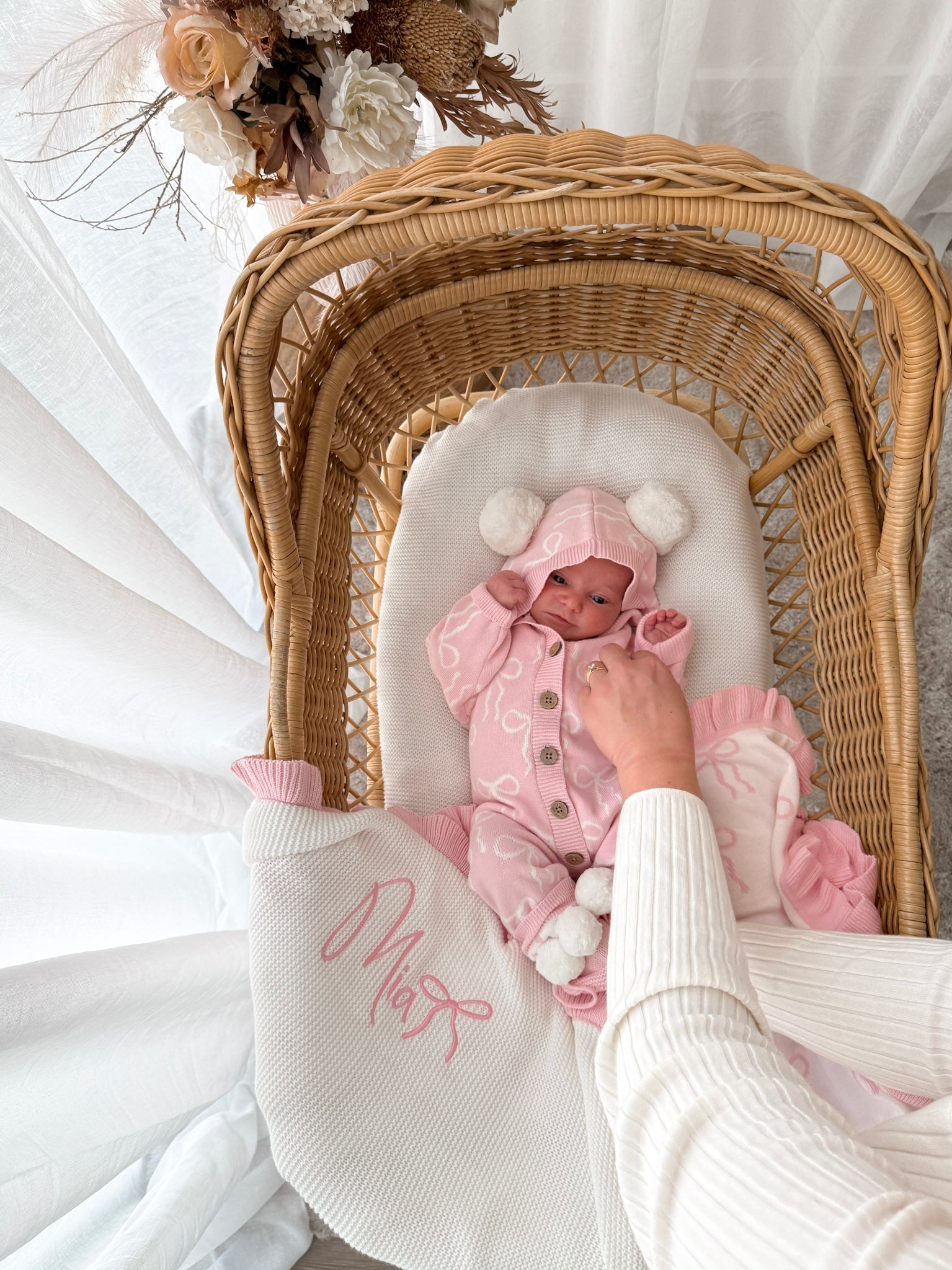 Baby in a pink hooded outfit with pom poms lying in a wicker crib with a white blanket. Decorative dried flowers in the background.