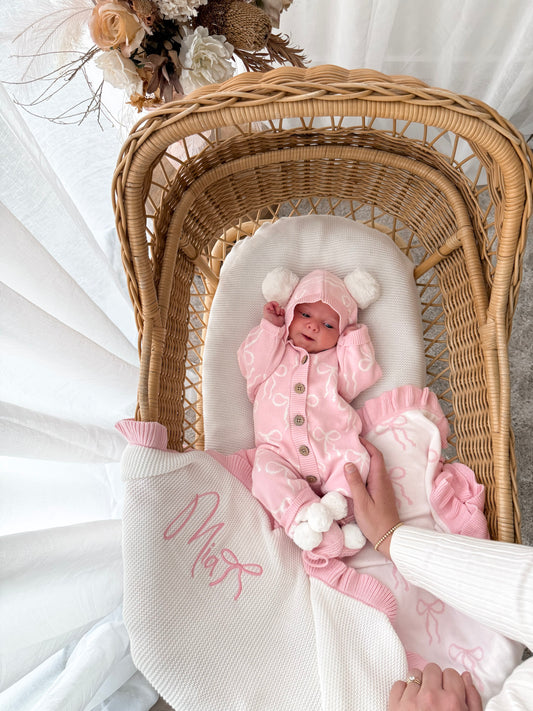 Baby in a pink hooded outfit with pom poms lying in a wicker crib with a white blanket. Decorative dried flowers in the background.