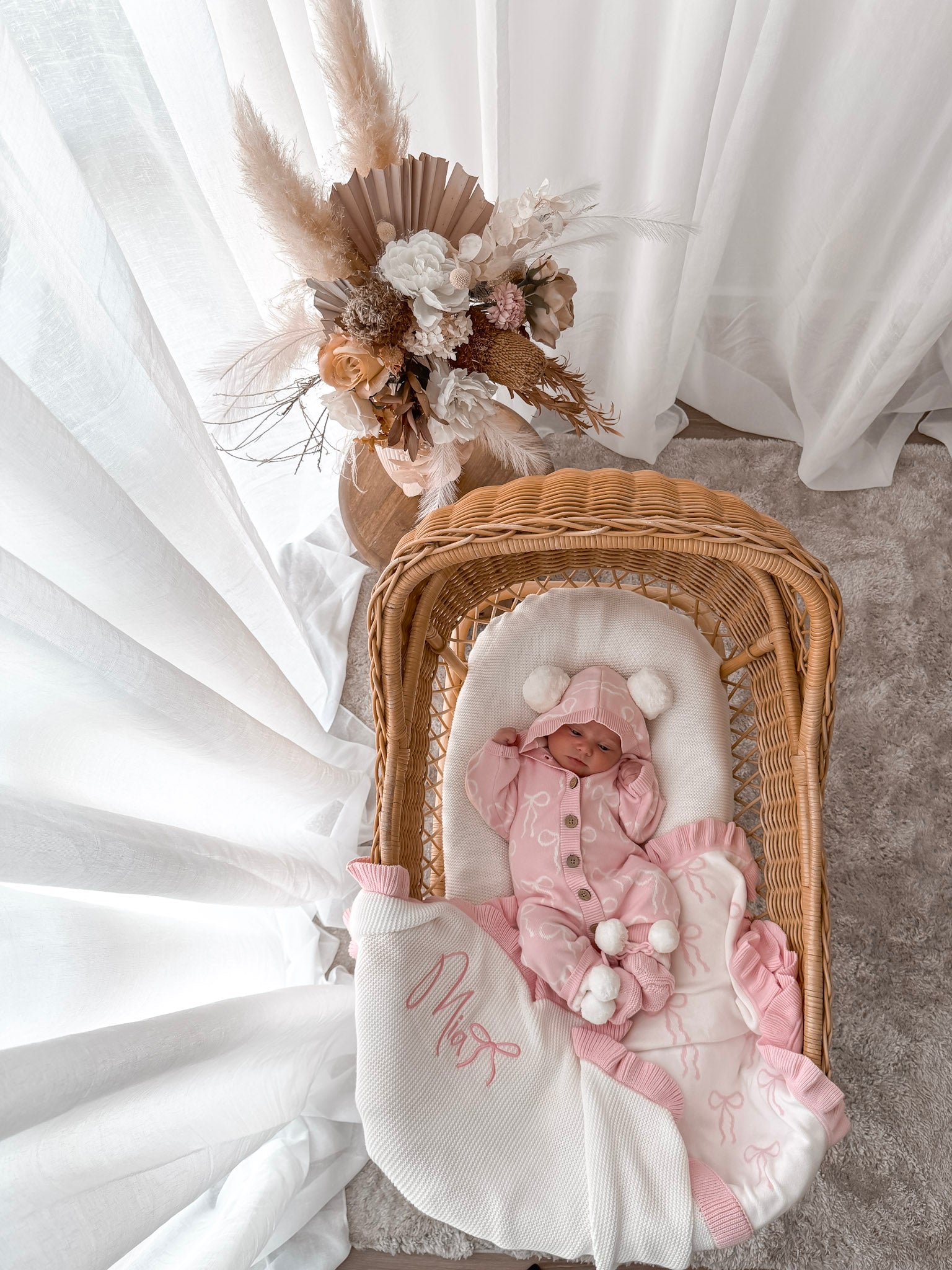 Baby in a pink hooded outfit with pom poms lying in a wicker crib with a white blanket. Decorative dried flowers in the background.