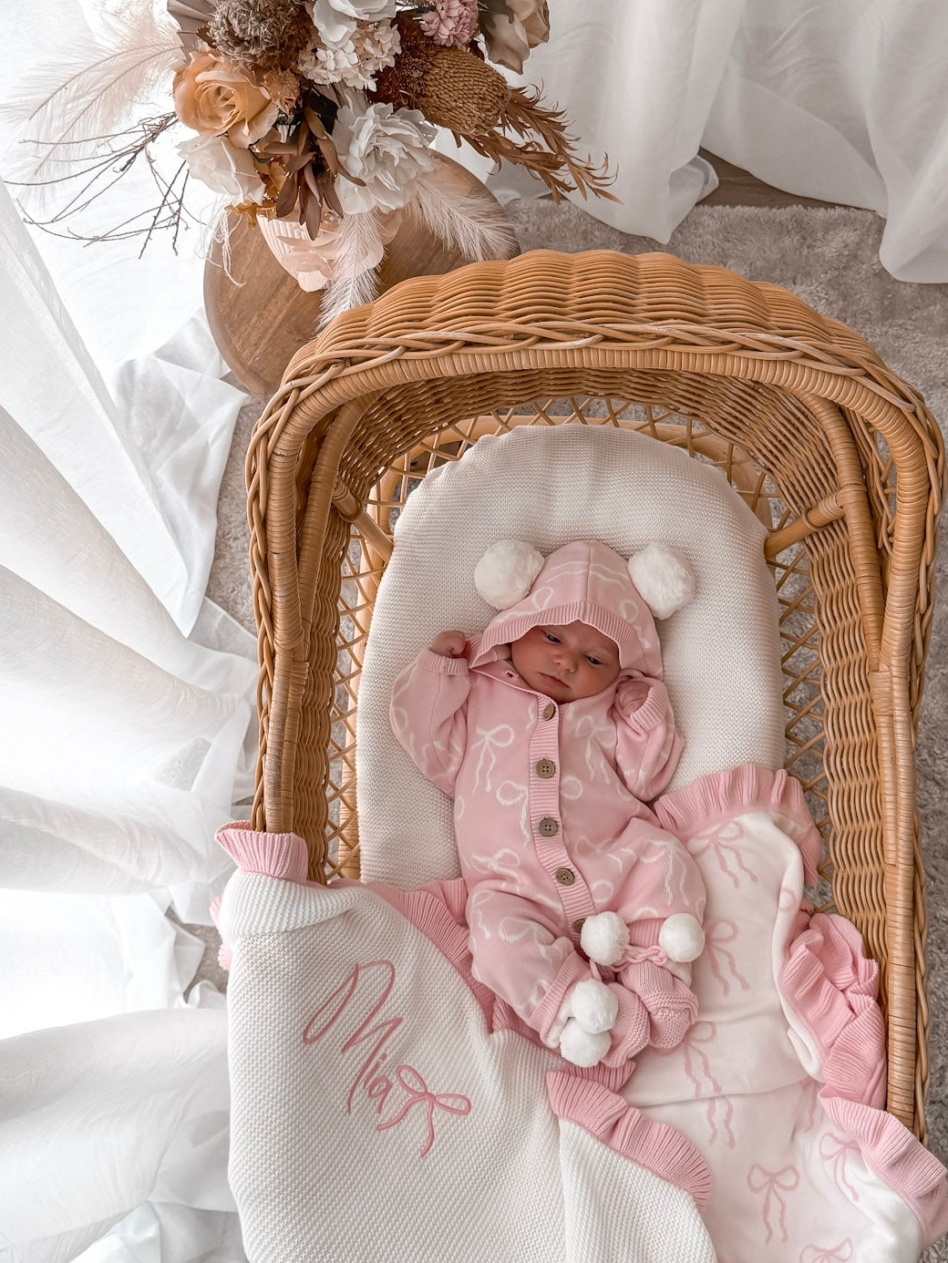 Baby in a pink hooded outfit with pom poms lying in a wicker crib with a white blanket. Decorative dried flowers in the background.