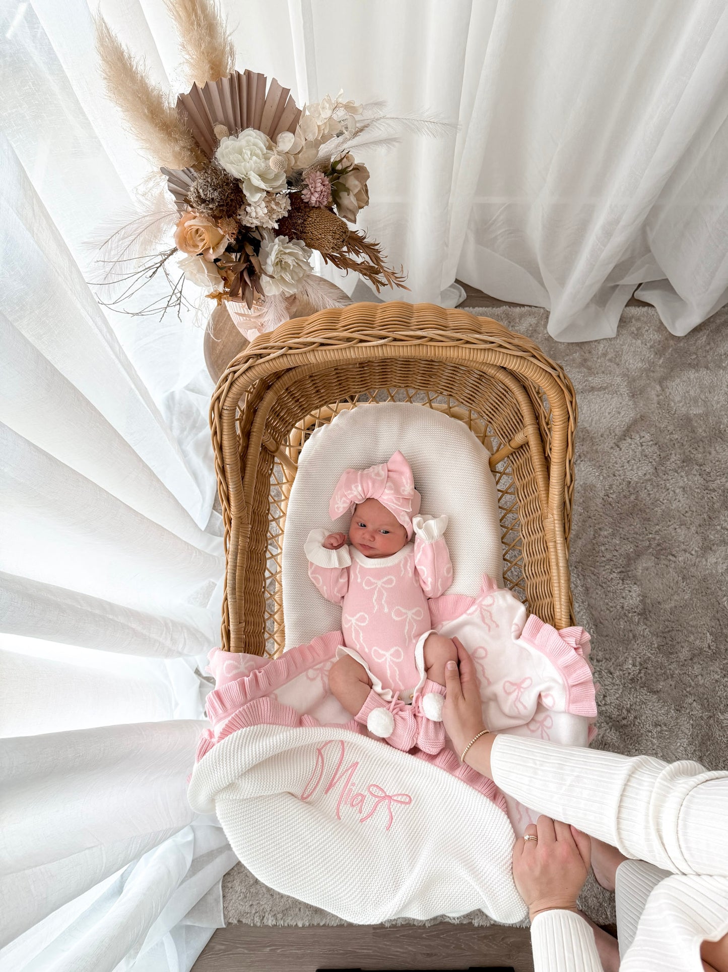 Newborn baby in a pink outfit lying in a wicker crib with decorative flowers on a white curtain background.