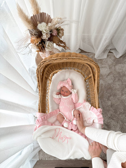 Newborn baby in a pink outfit lying in a wicker crib with decorative flowers on a white curtain background.