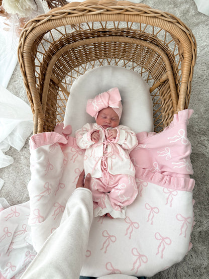 Baby in cardigan, pink and white pants, on a bow print blanket, in a wicker basket with dried florals in the background and sheer curtains surrounding two sides. 