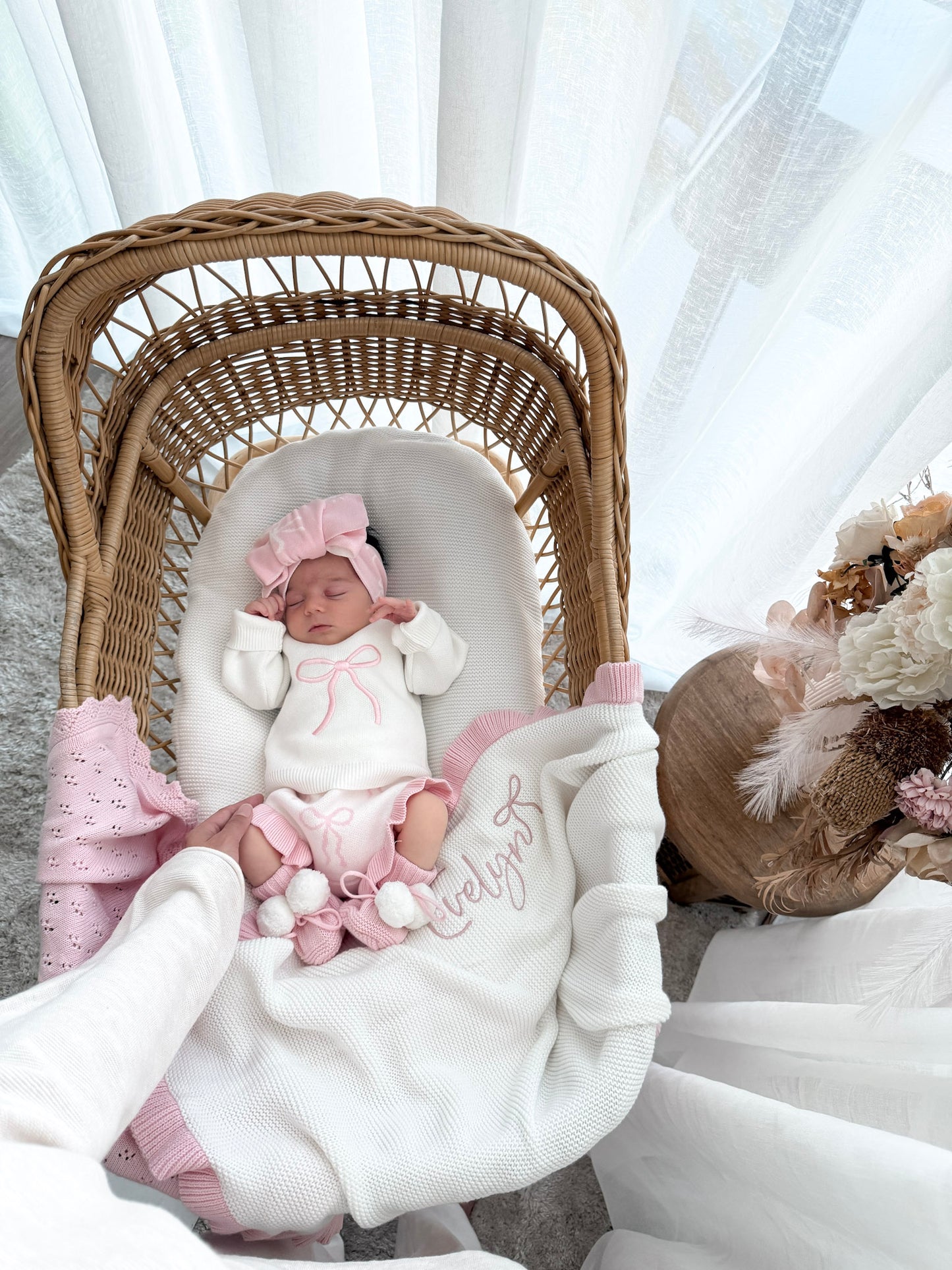 Newborn wearing a white jumper with a pink bow, bloomer bottoms with bow pattern and frills, bow pattern top knot, with a personalised whtie and pink blanket, in a wicker basket, sheer curtains and decorative dried florals.