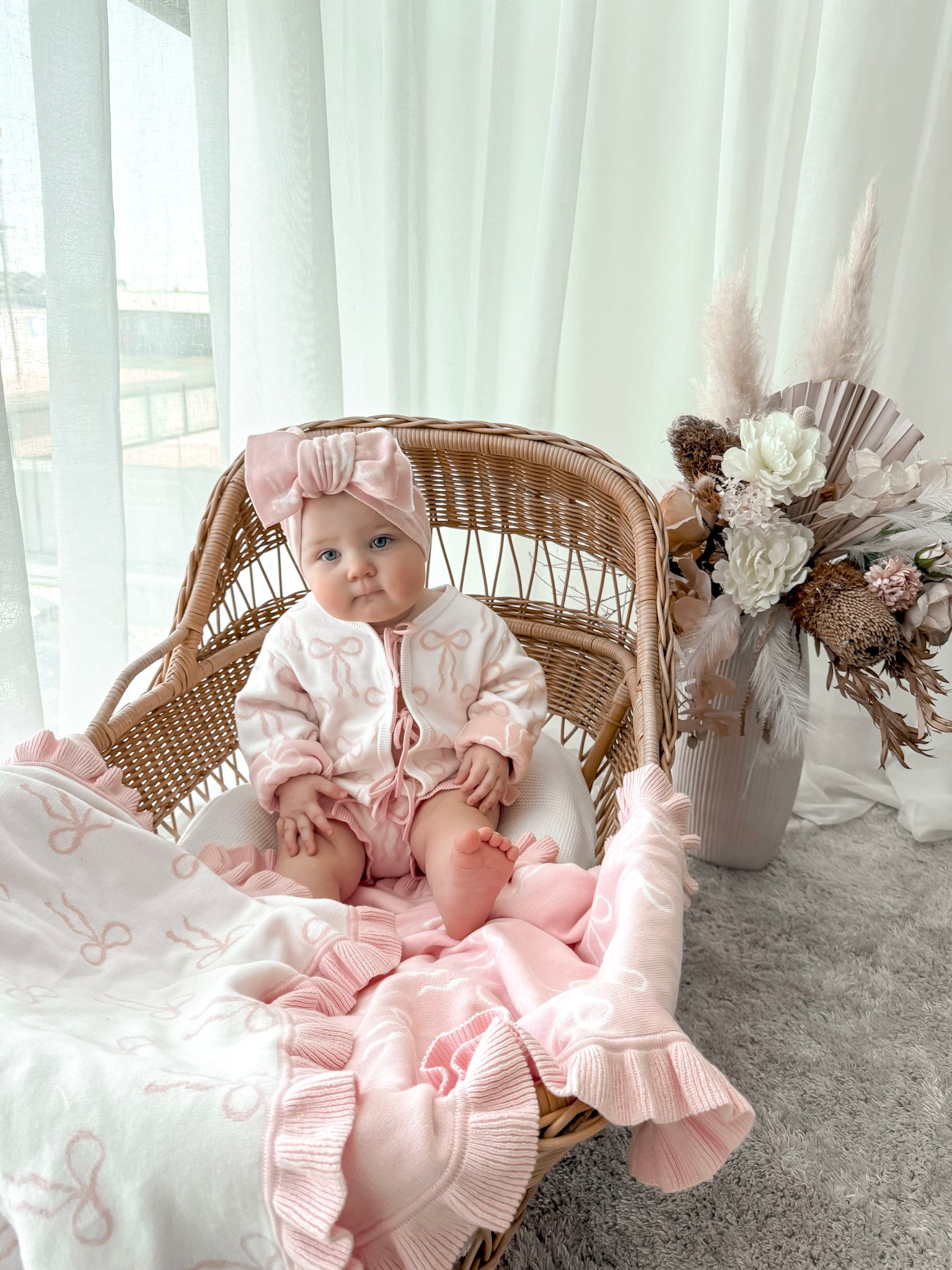 Baby in pink outfit sitting in a wicker chair with a floral arrangement in the background