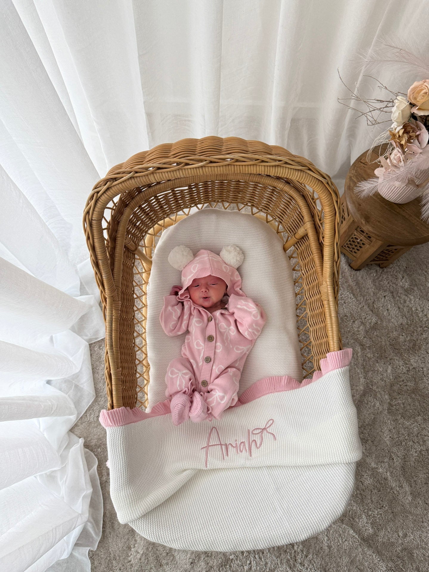 Baby in a pink hooded outfit with pom poms lying in a wicker crib with a white blanket. Decorative dried flowers in the background.