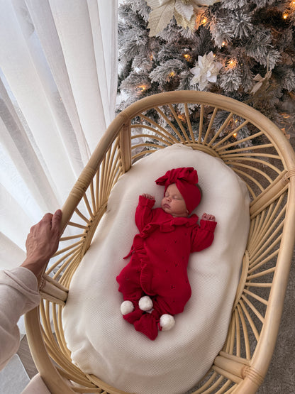 A newborn baby wearing a red knit romper and matching hat lies on a cream cushion inside a woven rattan bassinet. A hand holds the bassinet frame on the left, with a white curtain and a decorated Christmas tree in the background.