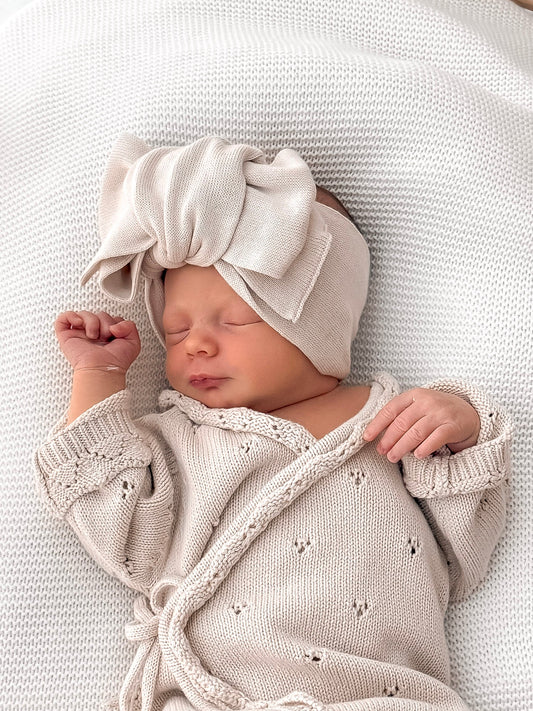 Newborn baby in a beige outfit and headband lying in a wicker basket with a white cushion.