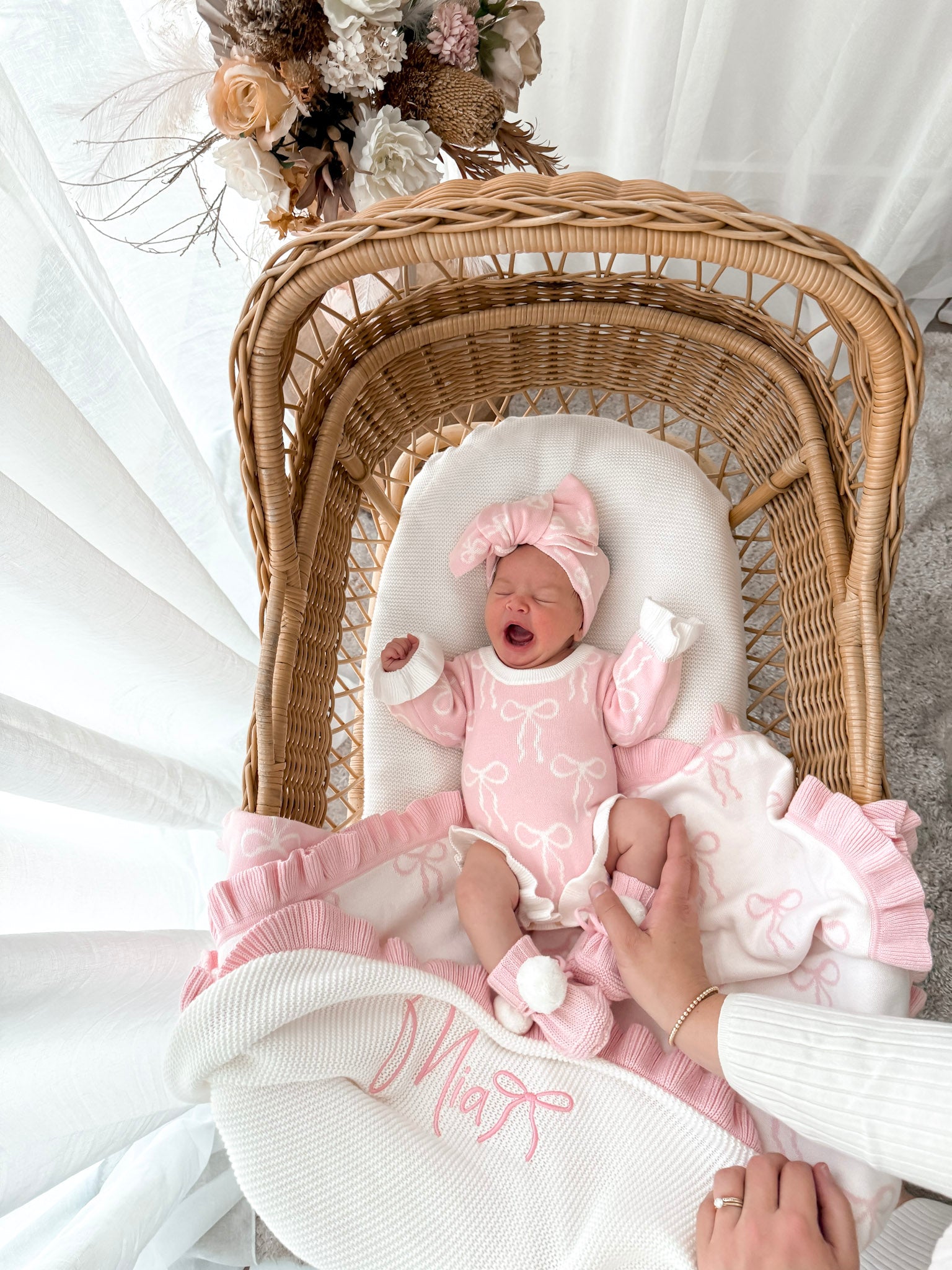Newborn baby in a pink outfit lying in a wicker crib with a white blanket yawning. 