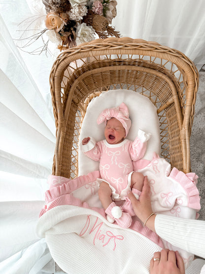 Newborn baby in a pink outfit lying in a wicker crib with a white blanket yawning. 