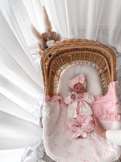 Newborn baby in a bow print cardigan, that ties in the front,bow print flared pants, both in pink & white, with a matching top knot. Baby is in a wicker backet, decorative dried florals behind. 