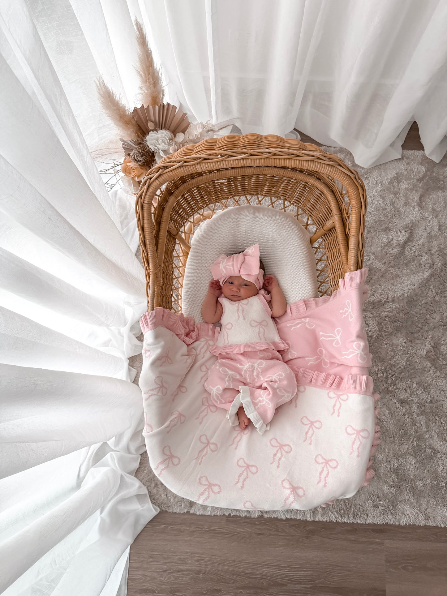 Baby in white and pink singlet top, pink and white pants, on a bow print blanket, in a wicker basket with dried florals in the background and sheer curtains surrounding two sides. 