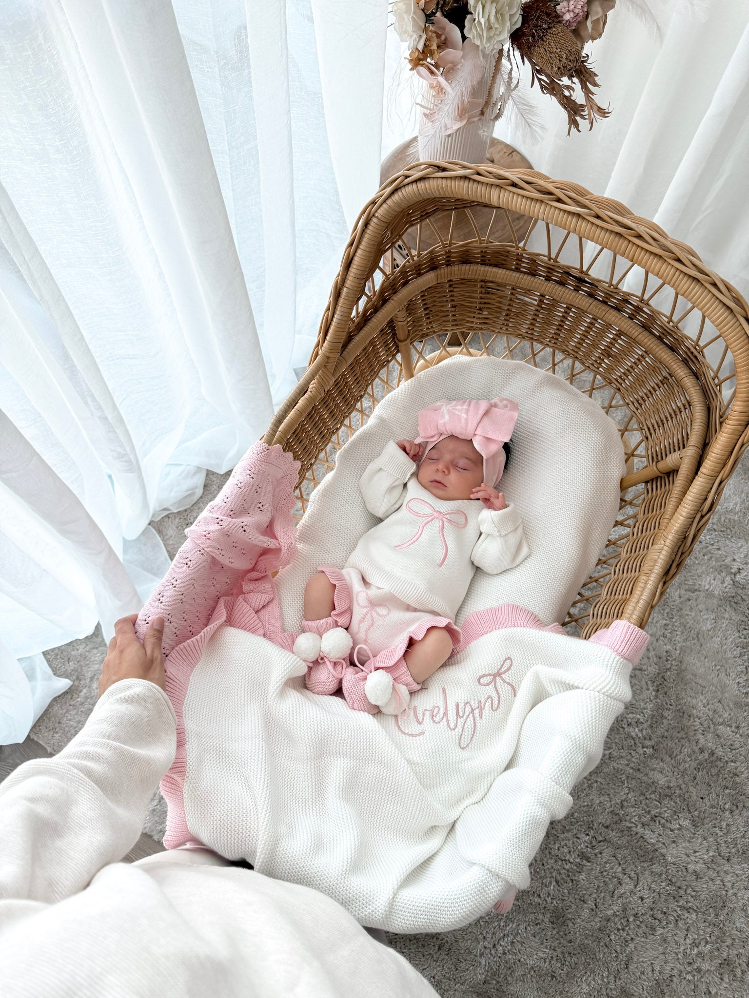 Newborn wearing a white jumper with a pink bow, bloomer bottoms with bow pattern and frills, bow pattern top knot, with a personalised whtie and pink blanket, in a wicker basket, sheer curtains and decorative dried florals. 