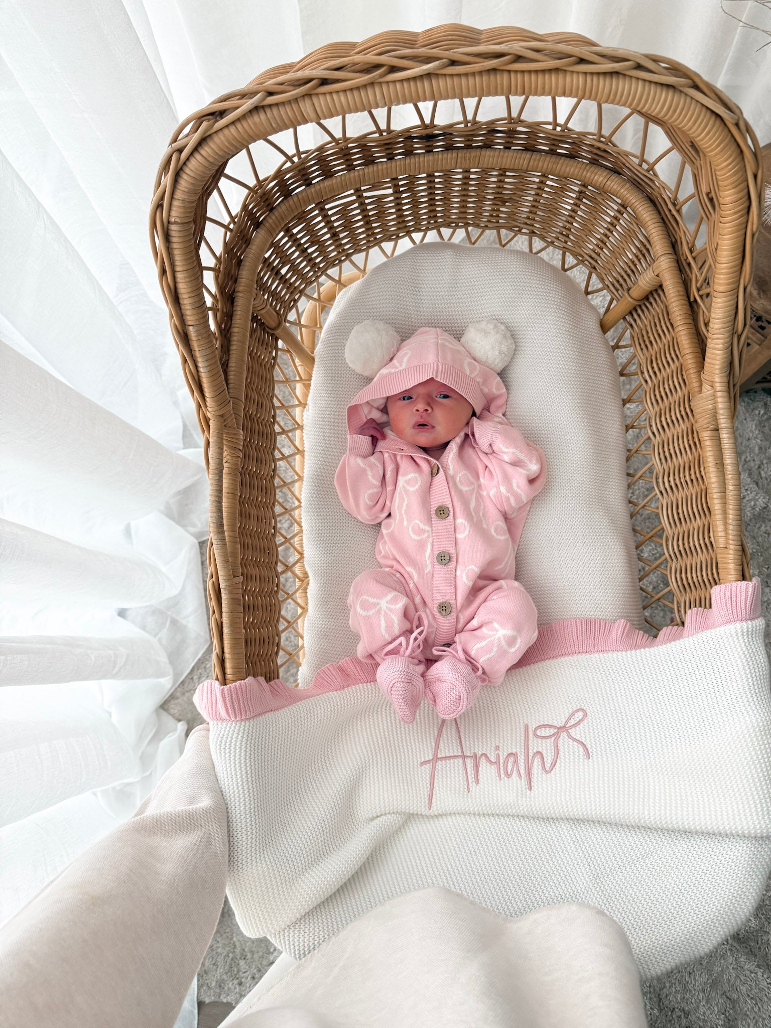 Baby in a pink hooded outfit with pom poms lying in a wicker crib with a white blanket. 