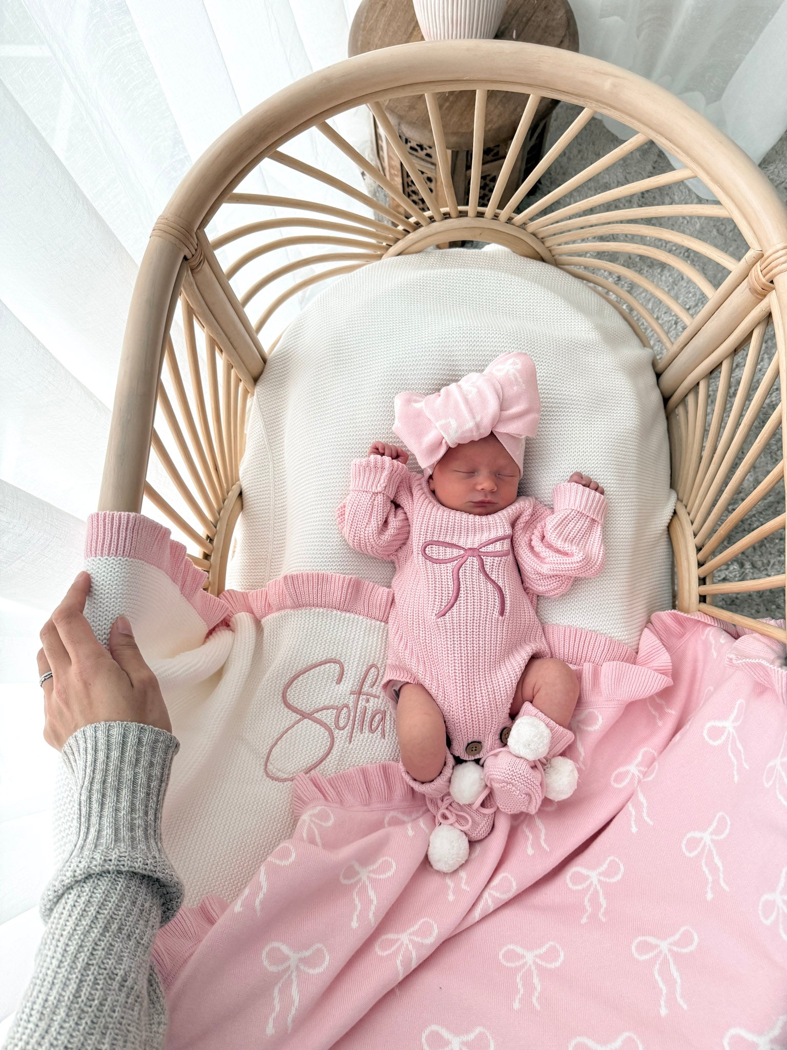 Newborn baby in a pink outfit with a bow, lying on a white blanket in a wicker chair.