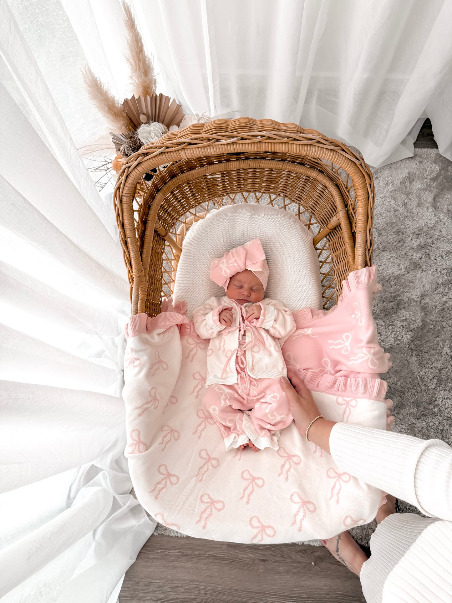 Baby in white and pink cardigan, pink and white pants, on a bow print blanket, in a wicker basket with dried florals in the background and sheer curtains surrounding two sides. 