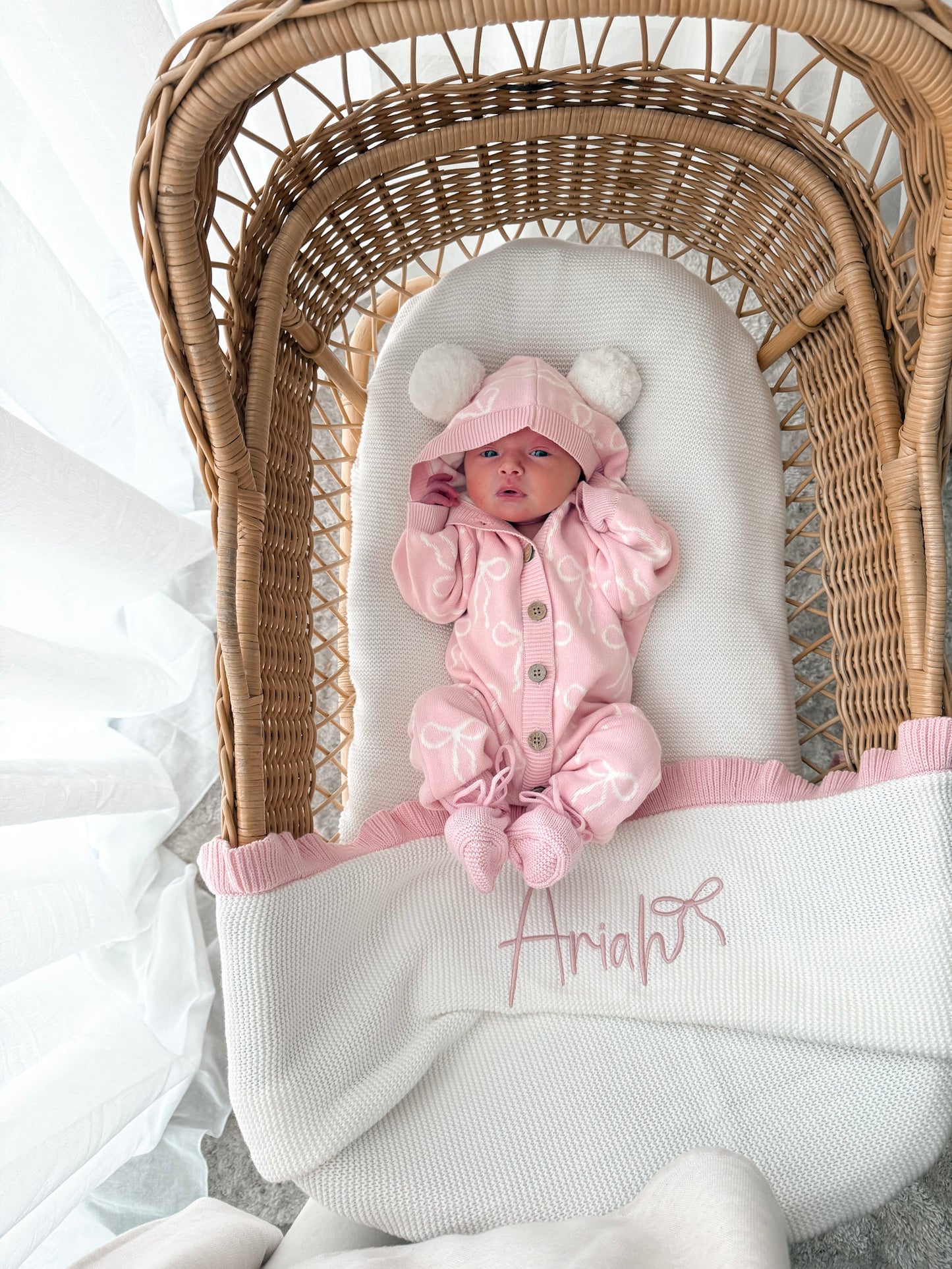 Baby in a pink hooded outfit with pom poms lying in a wicker crib with a white blanket. Decorative dried flowers in the background.