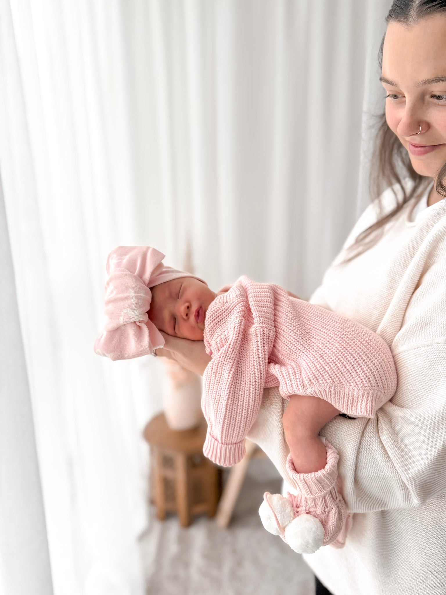 Woman holding a baby wrapped in pink clothing against a white curtain background