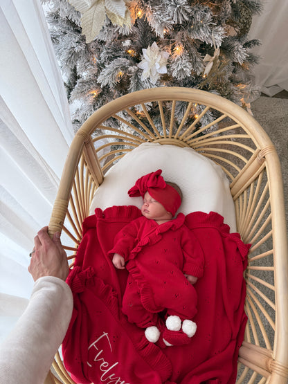 A baby wearing a red ruffled one-piece outfit and matching bow headband lies in a rattan baby chair with a white blanket, with a decorated Christmas tree in the background.
