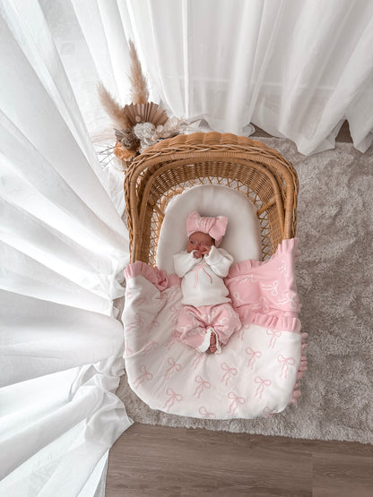 Baby in white jumper with a pink bow, pink and white pants, on a bow print blanket, in a wicker basket with dried florals in the background and sheer curtains surrounding two sides. 