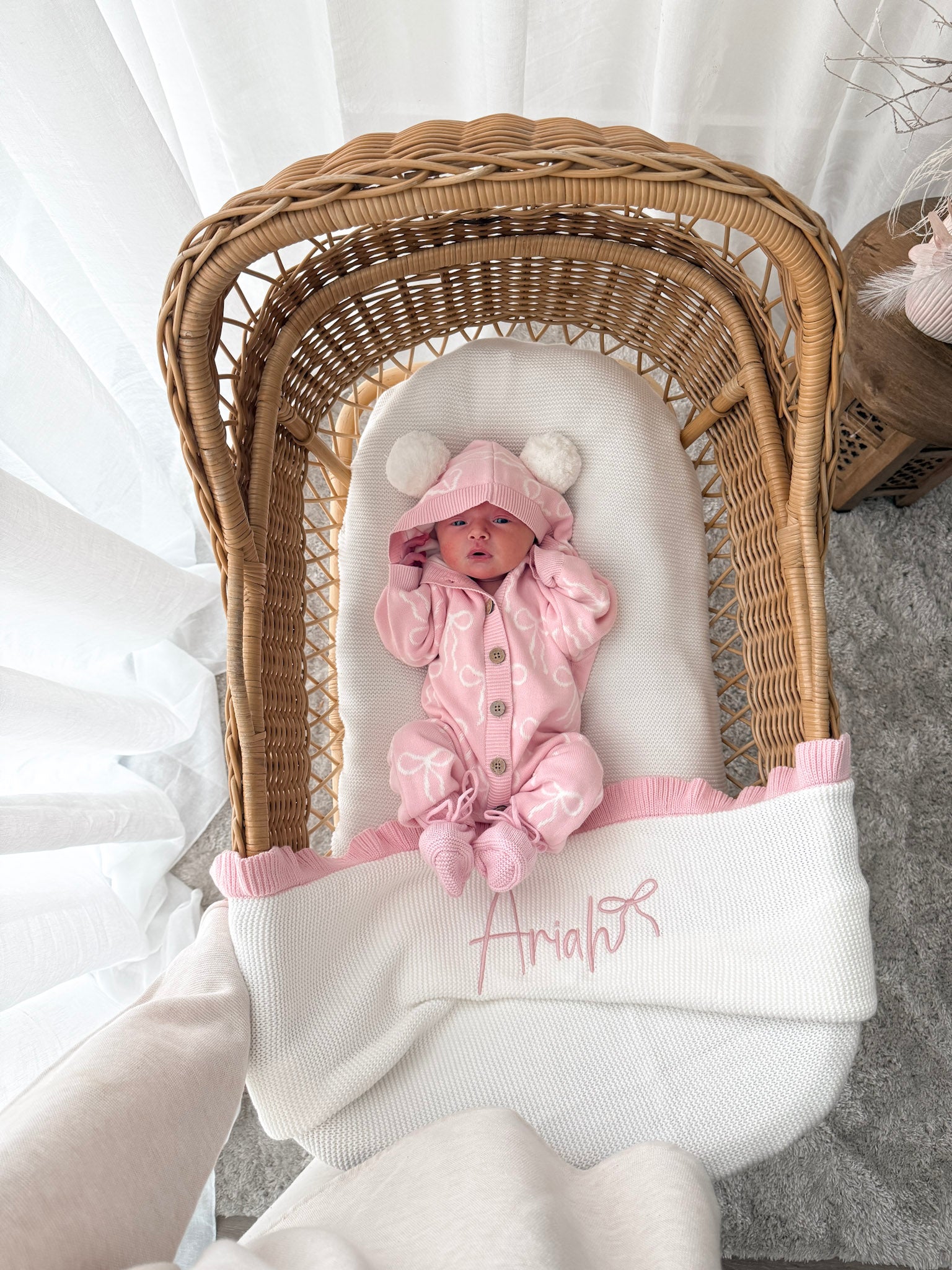 Baby in a pink hooded outfit with pom poms lying in a wicker crib with a white blanket. Decorative dried flowers in the background.