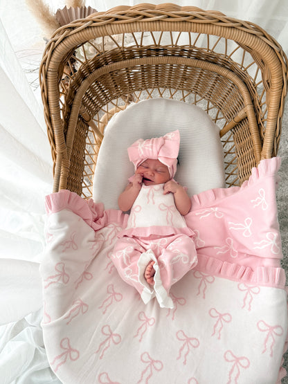 Baby in white and pink singlet top, pink and white pants, on a bow print blanket, in a wicker basket with dried florals in the background and sheer curtains surrounding two sides. 