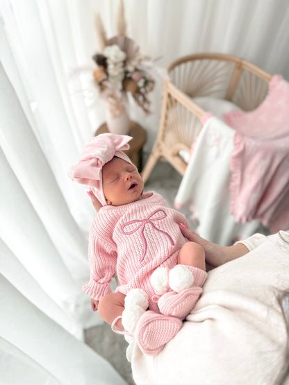 Newborn baby in a pink outfit with a matching hat, surrounded by soft textures and neutral colors.