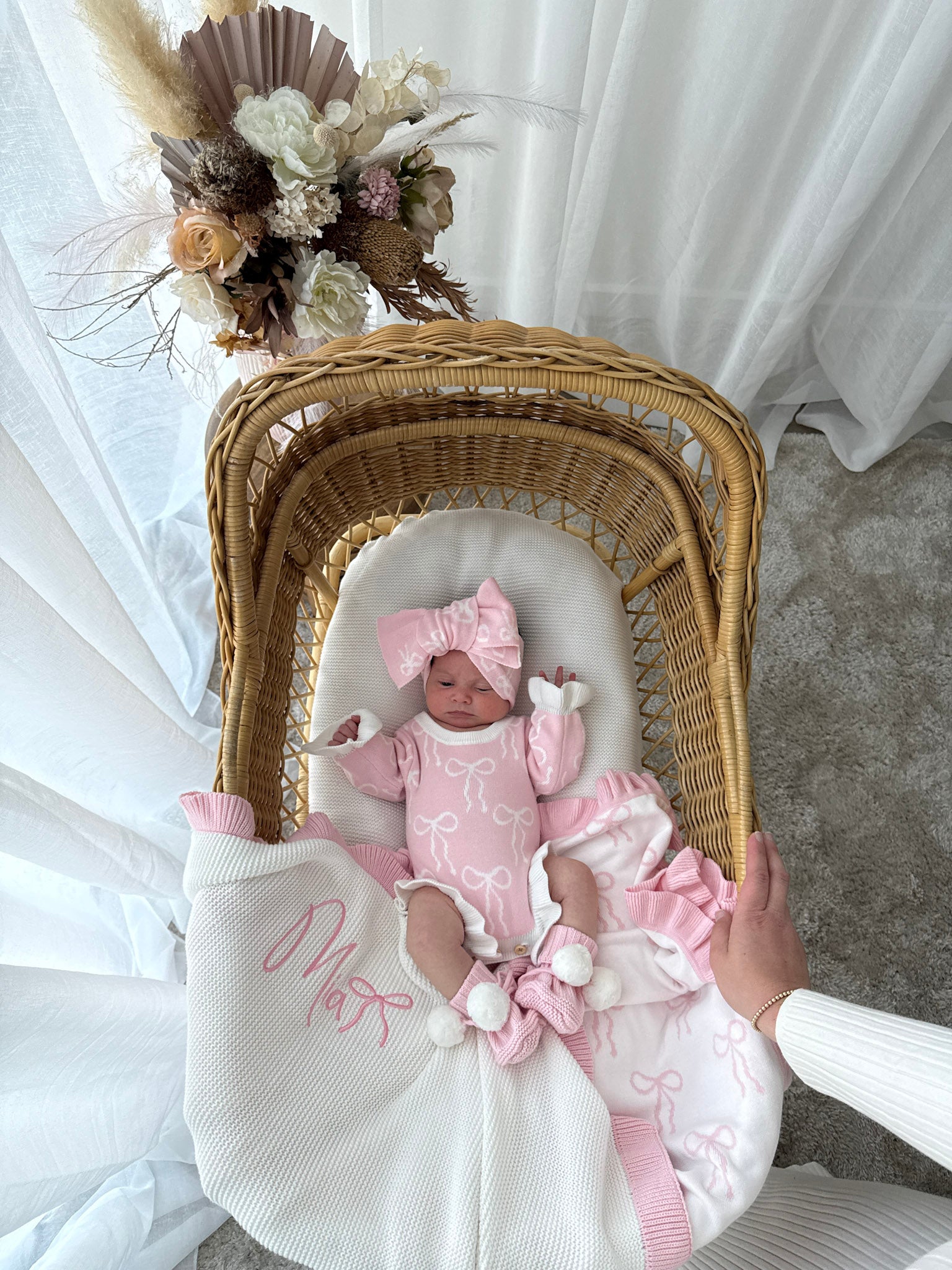 Baby doll in a pink outfit with a matching bow, lying on a white blanket next to a wicker basket.
