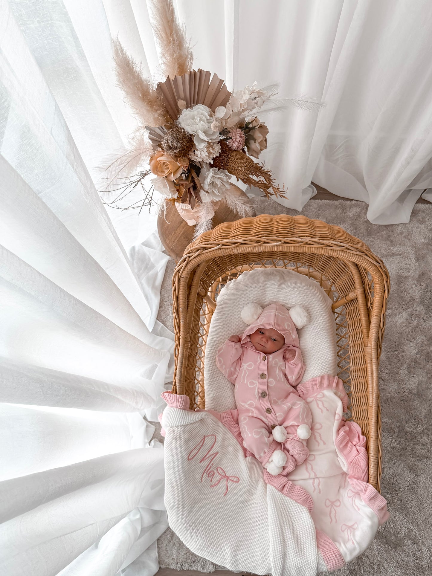 Baby in a pink hooded outfit with pom poms lying in a wicker crib with a white blanket. Decorative dried flowers in the background.