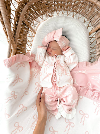 Newborn baby in a bow print cardigan, that ties in the front,bow print flared pants, both in pink & white, with a matching top knot. Baby is in a wicker basket.