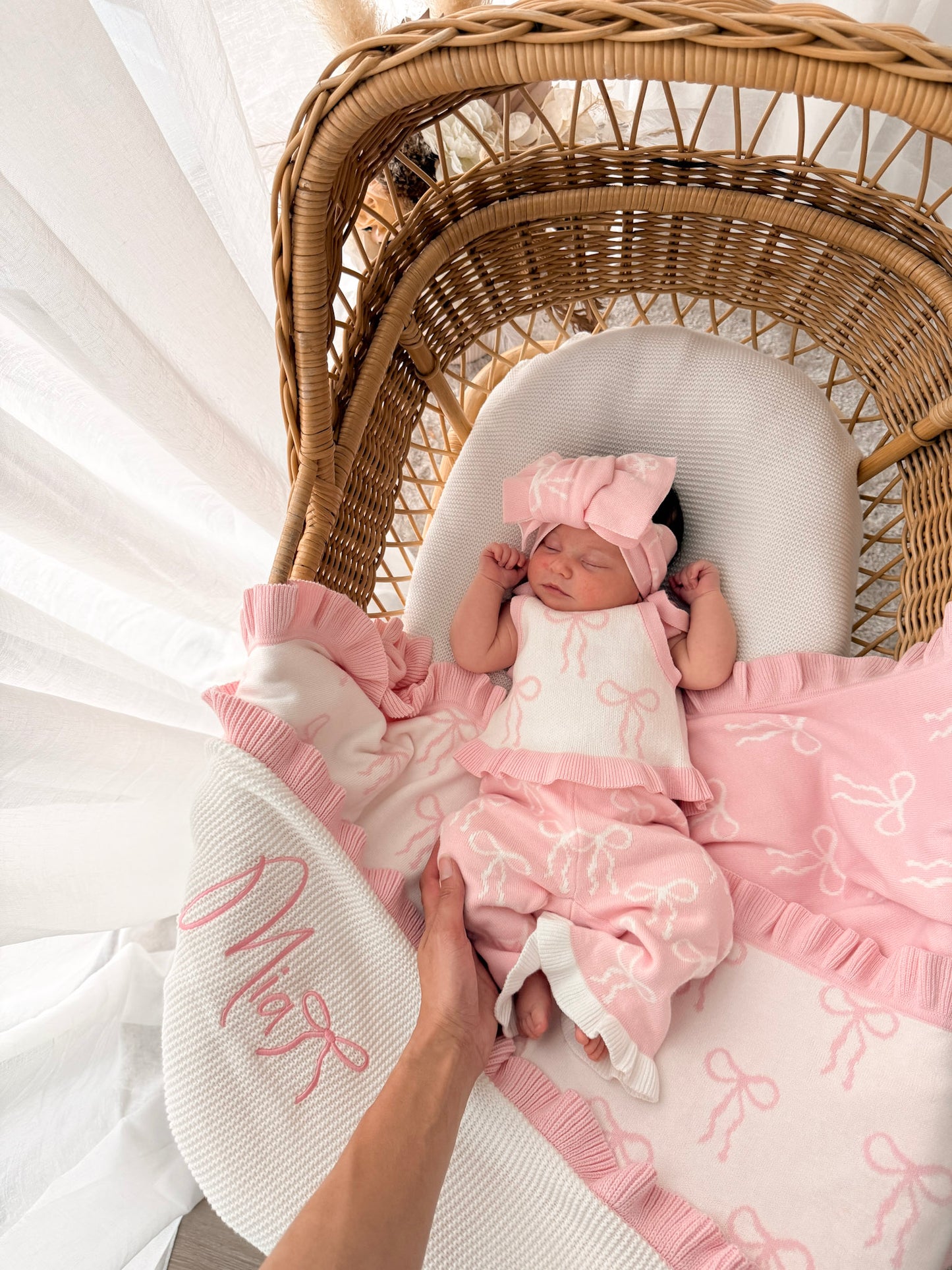 Baby in white and pink singlet top, pink and white pants, on a bow print blanket, in a wicker basket with dried florals in the background and sheer curtains surrounding two sides. 
