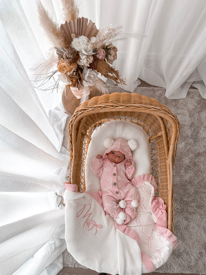 Baby in a pink hooded outfit with pom poms lying in a wicker crib with a white blanket. Decorative dried flowers in the background.