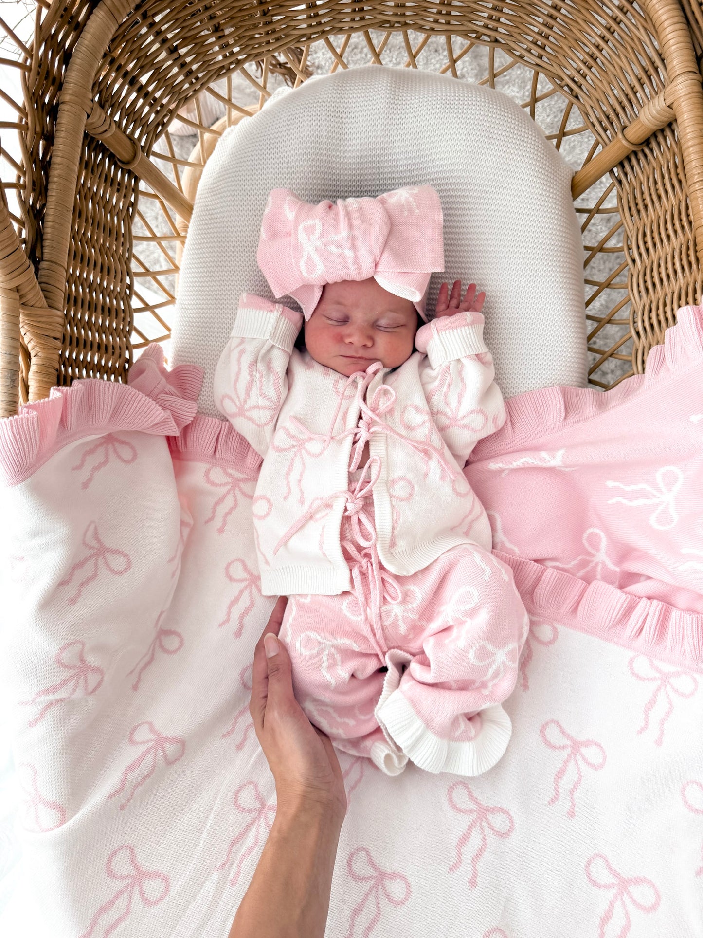 Newborn baby in a bow print cardigan, that ties in the front,bow print flared pants, both in pink & white, with a matching top knot. Baby is in a wicker basket.