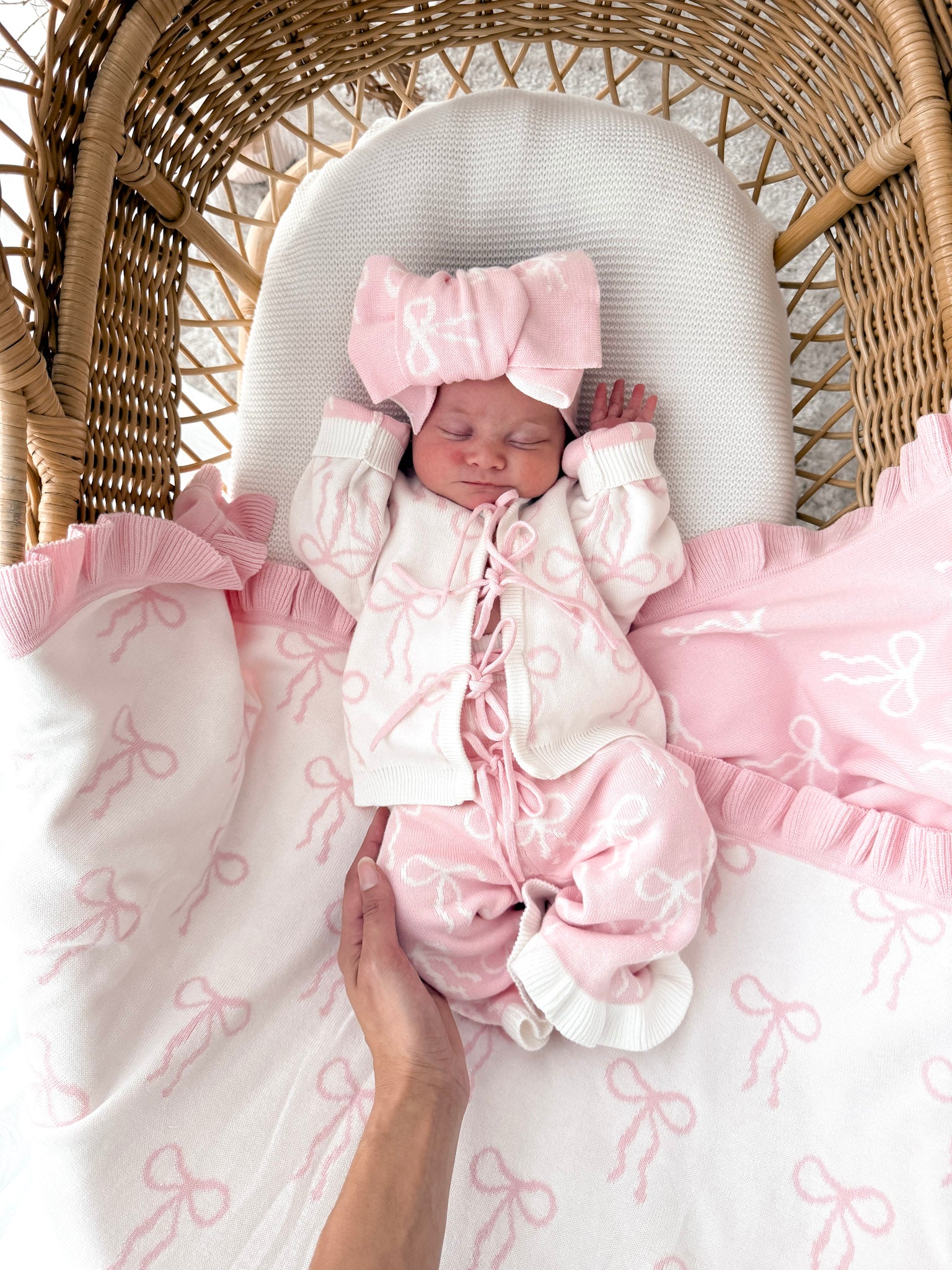 Newborn baby in a bow print cardigan, that ties in the front,bow print flared pants, both in pink & white, with a matching top knot. Baby is in a wicker basket.