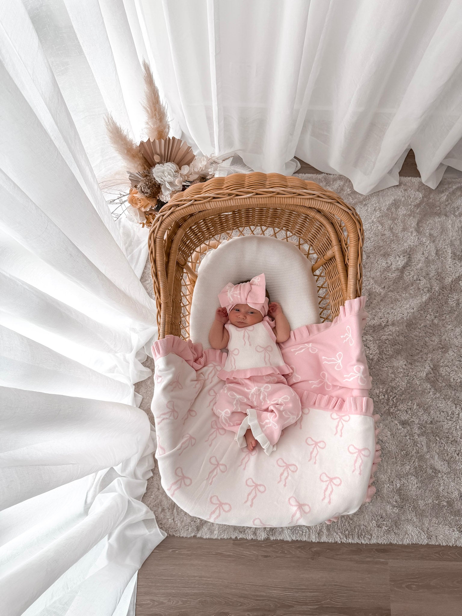 Baby in white and pink singlet top, pink and white pants, on a bow print blanket, in a wicker basket with dried florals in the background and sheer curtains surrounding two sides. 