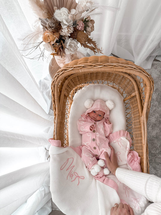 Baby in a pink hooded outfit with pom poms lying in a wicker crib with a white blanket. Decorative dried flowers in the background.