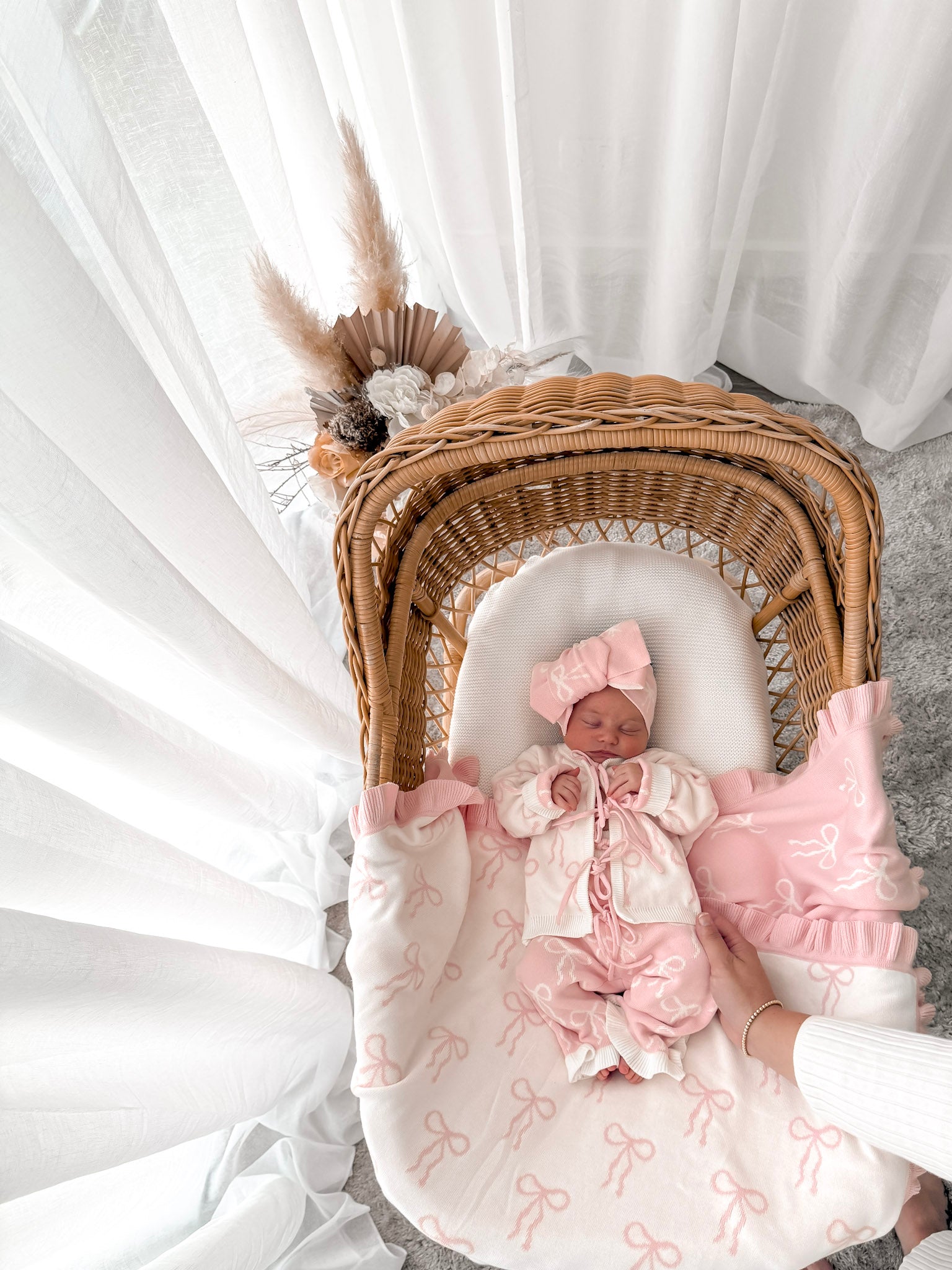 Newborn baby in a bow print cardigan, that ties in the front,bow print flared pants, both in pink & white, with a matching top knot. Baby is in a wicker backet, decorative dried florals behind. 