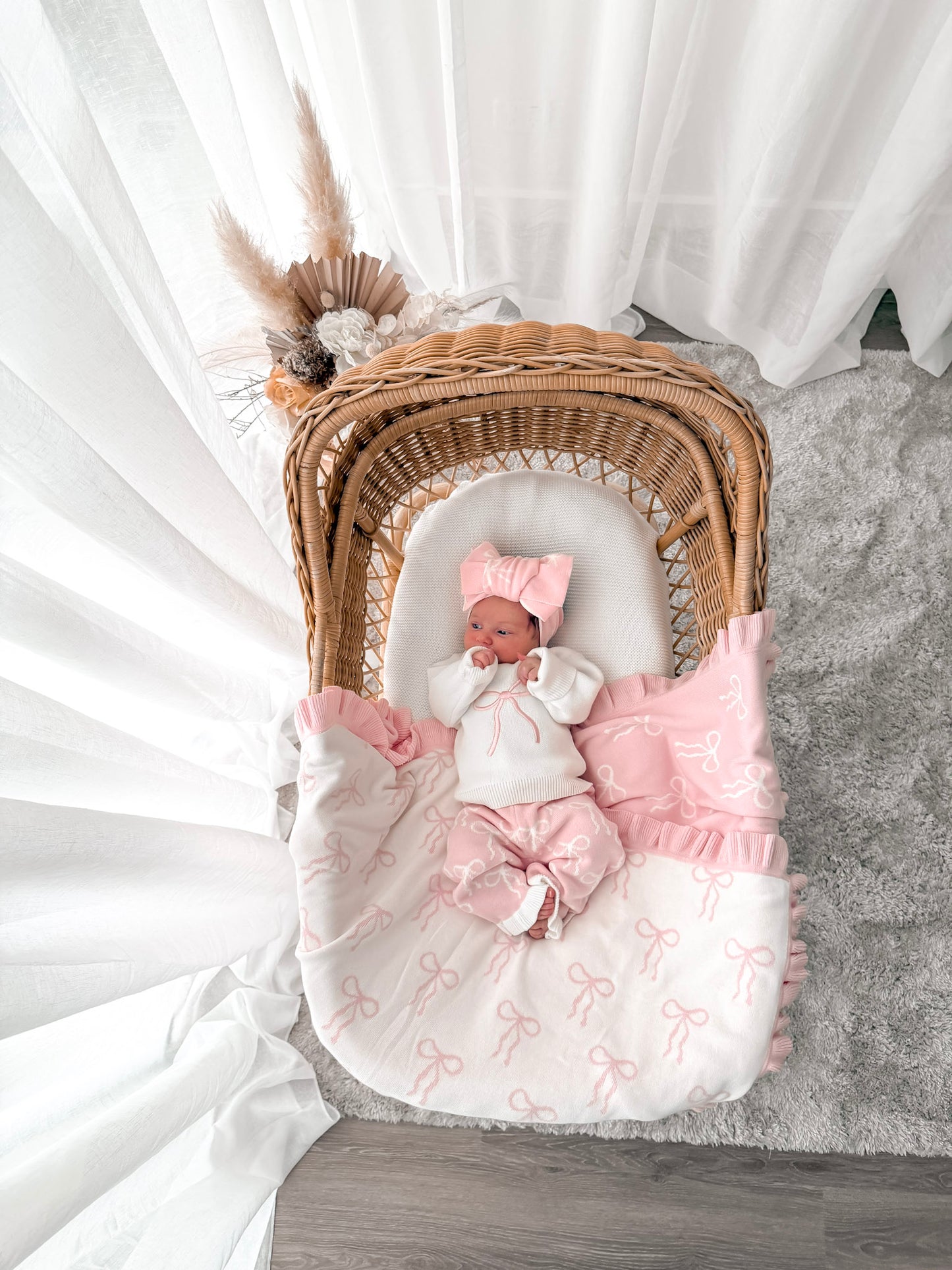 Baby in white and pink jumper top, pink and white pants, on a bow print blanket, in a wicker basket with dried florals in the background and sheer curtains surrounding two sides. 