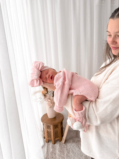 Woman holding a baby wrapped in pink clothing and headband against a white curtain background