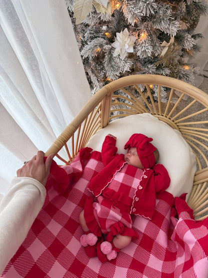 A baby wearing a red knit holiday outfit and matching hat rests in a rattan bassinet draped with a red and pink buffalo plaid blanket, with a white curtain and a decorated Christmas tree in the background.