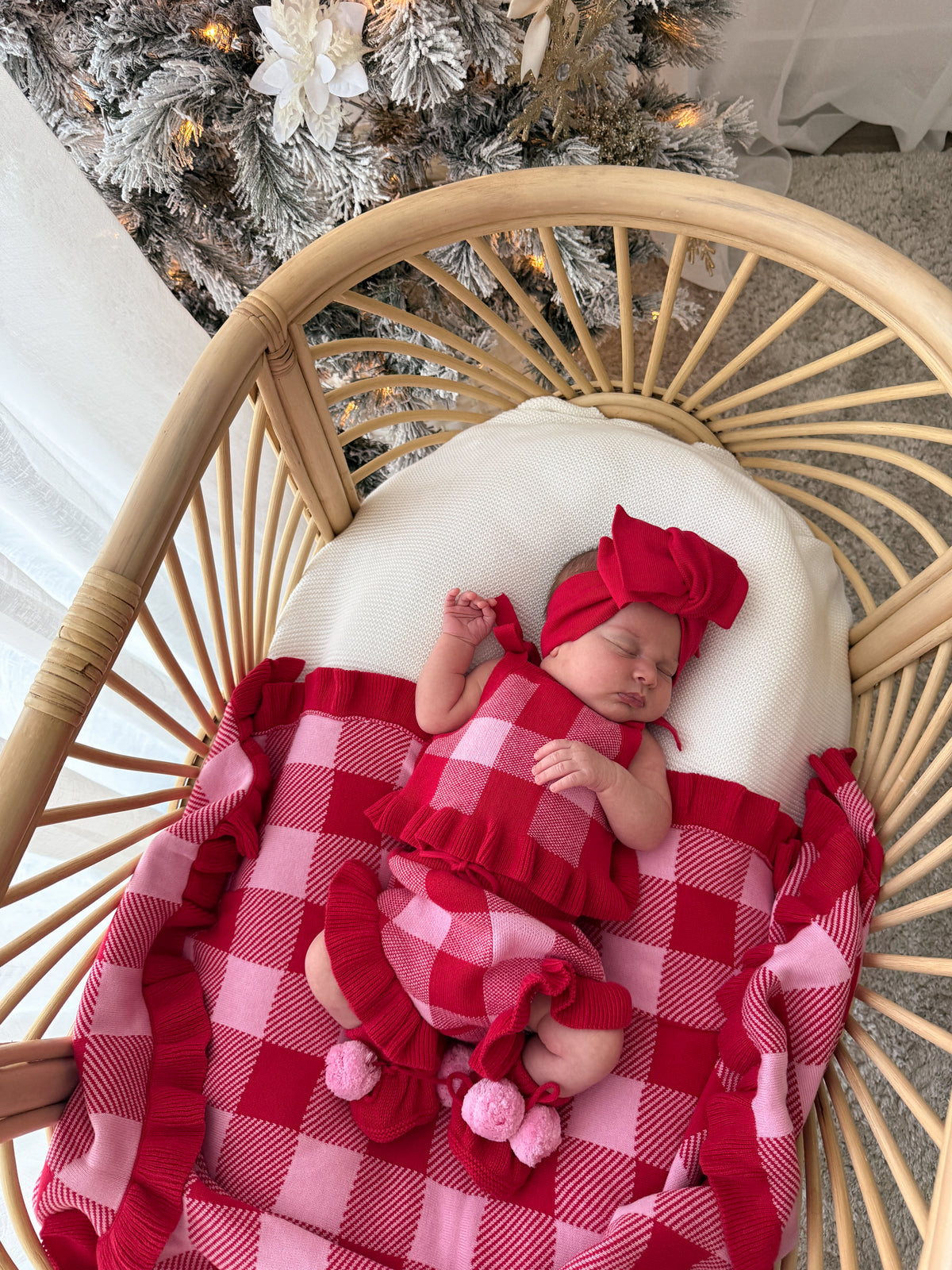 A newborn baby wearing a red and pink gingham outfit with a matching headband, sleeping in a rattan baby chair with a white cushion and a red and pink gingham blanket. A decorated Christmas tree is visible in the background.